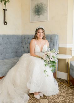 a woman in a wedding dress sitting on a blue couch holding a bouquet and smiling at the camera