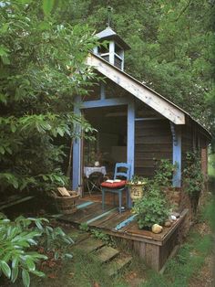 a small wooden cabin in the woods surrounded by greenery and trees, with a table and chairs outside