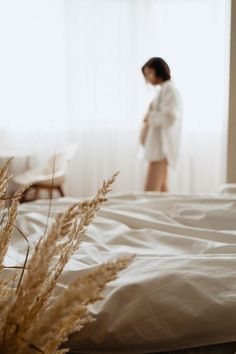 a woman standing in front of a window next to a bed covered in white sheets