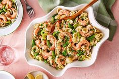 a bowl of pasta with shrimp and asparagus on a pink surface next to two plates