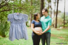 a pregnant woman and man standing next to each other in the grass with a baby's dress hanging from a tree