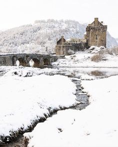 an old castle sits on top of a snowy hill next to a small river and bridge