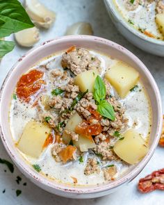 two bowls of soup with meat, potatoes and tomatoes on the side next to basil leaves