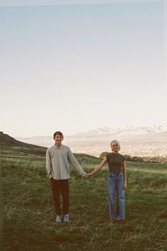 a man and woman holding hands while standing on top of a lush green field with mountains in the background