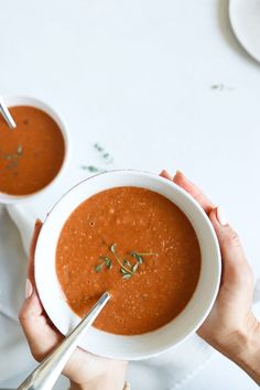 two hands holding bowls of tomato soup on a white table with silverware and napkins