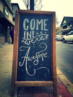 a chalkboard sign on the side of a street that says come in, we're awesome