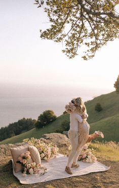 two people are hugging on a blanket in the grass with flowers and pillows around them