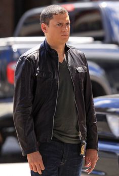 a man in black jacket and jeans standing on street next to parked cars with his hands in his pockets