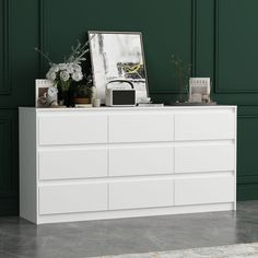 a white dresser with flowers and pictures on it next to a dark green painted wall