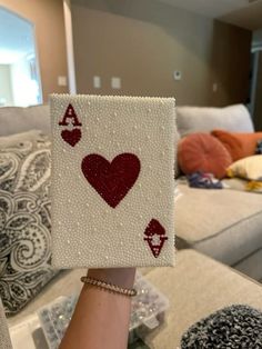 a hand holding up a playing card made out of beads and sequins in front of a couch