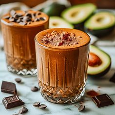 two glasses filled with chocolate pudding next to sliced avocado and chocolate chips on a table