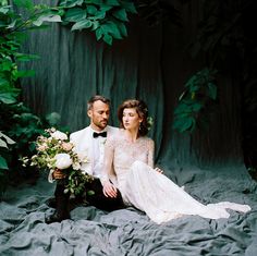 a bride and groom sitting on the ground in front of some greenery holding bouquets