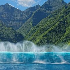 an ocean wave is breaking in front of some mountains and trees on the water's edge