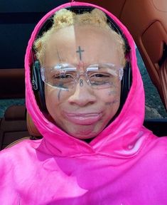 a woman wearing glasses and a pink hoodie has her face painted with cross on it