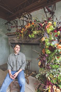 a man sitting on a stool in front of a fireplace with flowers hanging from it