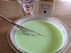 a bowl filled with green liquid next to a carton of milk and a whisk