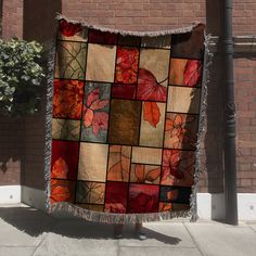 a woman is standing on the sidewalk holding up a quilt with leaves and flowers in it