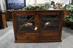 a wooden cabinet with glass doors on display in a room filled with plants and people