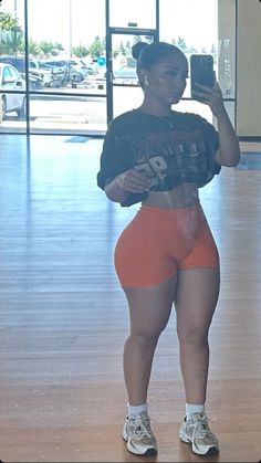 a woman taking a selfie with her cell phone in an empty gym room while wearing orange shorts