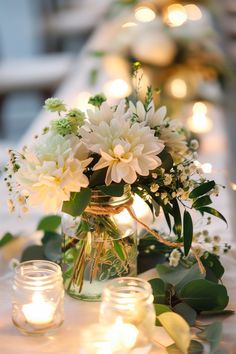 white flowers and greenery are in a mason jar on a table with lit candles