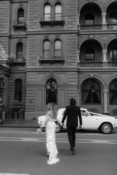 a bride and groom walking across the street in front of an old building with cars