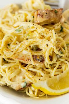 a white plate topped with pasta covered in chicken and lemon wedges next to a fork