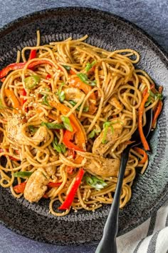 a black plate filled with noodles and chicken on top of a blue table cloth next to a fork