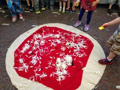 a group of children standing around a large red piece of cloth on the ground with white flowers all over it