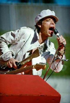 a man singing into a microphone while holding a guitar in his right hand and wearing a white hat