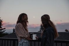 two women standing next to each other on a balcony holding coffee mugs in their hands