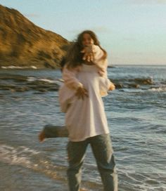 a woman walking along the beach carrying a baby in her arms and smiling at the camera