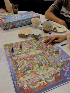 two people sitting at a table with a board game in front of them and other items on the table