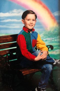 a young boy sitting on a wooden bench in front of a rainbow painted wall behind him