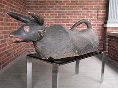 a wooden sculpture of a dog laying on top of a metal stand in front of a brick wall