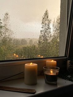 two candles sitting on a window sill in front of a rain soaked window with trees outside