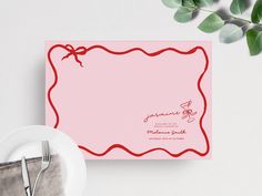 a pink card with a red ribbon on it next to a white plate and silverware