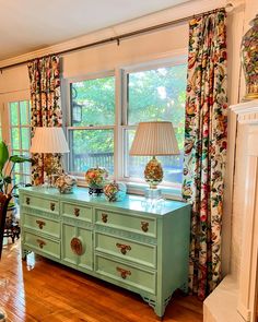 a green dresser with two lamps on top of it in front of a large window