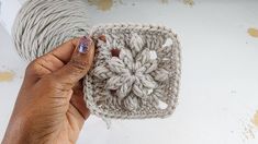 a hand holding up a crochet square on top of a white surface next to a ball of yarn