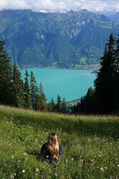 a woman sitting on top of a lush green hillside next to a lake in the mountains