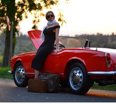 a woman standing on the hood of a red car with luggage in front of it