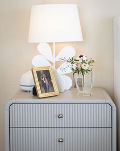 a white dresser topped with a lamp next to a vase filled with flowers and a framed photo