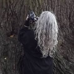 a woman with long white hair taking a photo in front of a tree while holding a camera