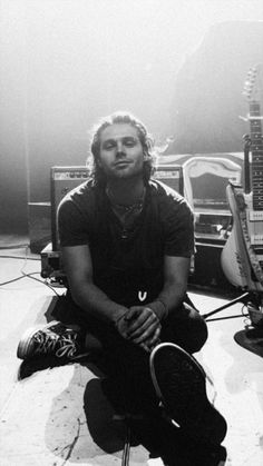 a man sitting on the ground with his guitar in front of him and other guitars behind him