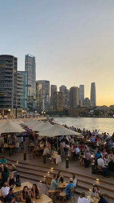 many people are sitting at tables near the water in front of tall buildings and skyscrapers