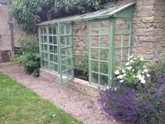 an old greenhouse is surrounded by flowers and greenery in the back yard with stone walls