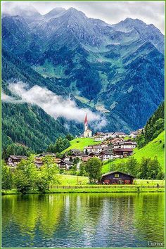 a lake with mountains in the background and houses on the shore, surrounded by green grass