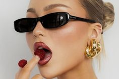 a woman wearing sunglasses and holding raspberries in her mouth with one hand on the lip