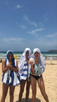 three women in bathing suits standing on the beach with towels around their necks and one holding a cell phone