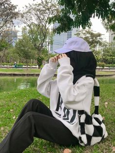 a person sitting on the ground in front of a pond wearing a white and black hoodie