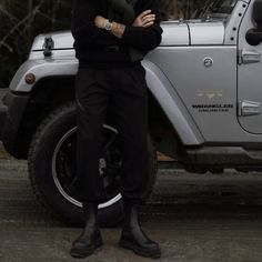 a man standing in front of a jeep with his arms crossed and wearing black boots